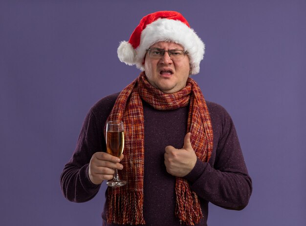 보라색 벽에 고립 된 엄지 손가락을 보여주는 샴페인 잔을 들고 목에 스카프와 안경과 산타 모자를 쓰고 자신감이 성인 남자