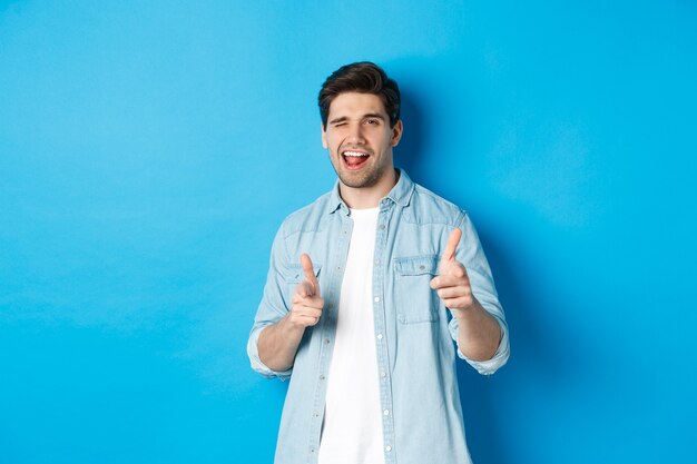 Confident adult man winking, pointing finger at you and winking, congratulating or praising person, standing against blue background