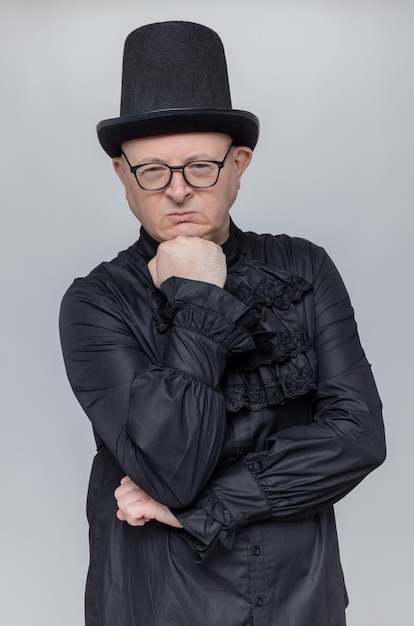 Free photo confident adult man with top hat and glasses in black gothic shirt putting fist on his chin and looking