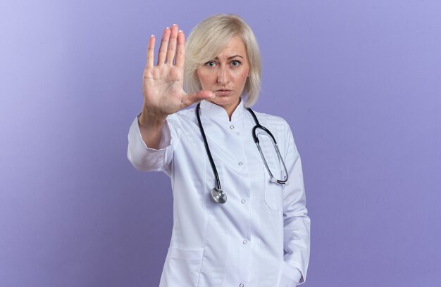 Confident adult slavic female doctor in medical robe with stethoscope gesturing stop hand sign isolated on purple background with copy space