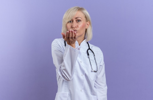 Confident adult slavic female doctor in medical robe with stethoscope sending kiss with hand isolated on purple wall with copy space