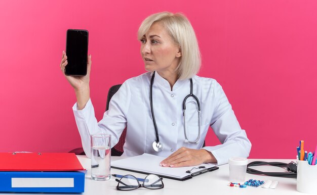 Confident adult slavic female doctor in medical robe with stethoscope sitting at desk with office tools holding and looking at phone isolated on pink background with copy space