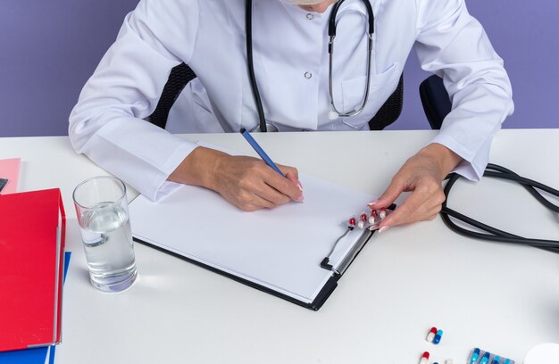 Confident adult slavic female doctor in medical robe with stethoscope sitting at desk with office tools writing on clipboard with pen isolated on purple background with copy space