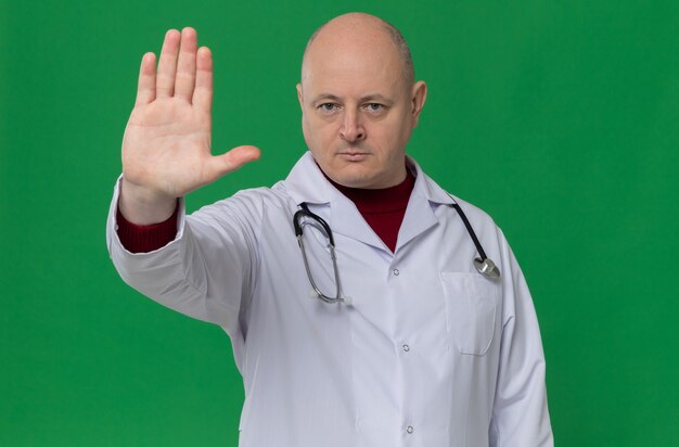 Confident adult slavic man in doctor uniform with stethoscope gesturing stop hand sign 