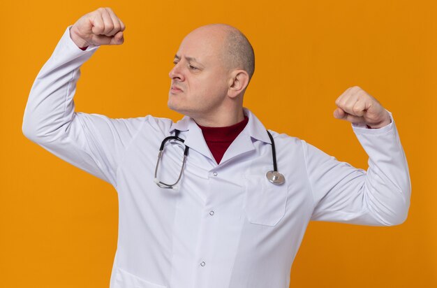 Уверенный взрослый славянский мужчина в медицинской форме со стетоскопом, напрягающий бицепс и смотрящий в сторону