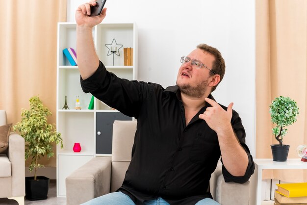 Confident adult slavic man in optical glasses sits on armchair pointing at himself and looking at phone taking selfie inside living room