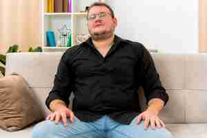 Free photo confident adult slavic man in optical glasses sits on armchair putting hands on legs inside living room