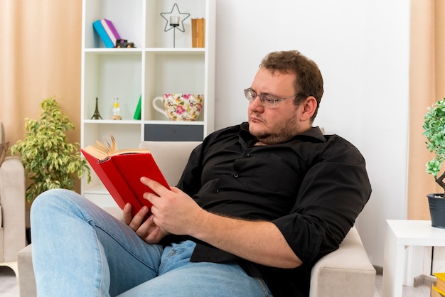 Free photo confident adult slavic man in optical glasses sits on armchair reading book inside living room