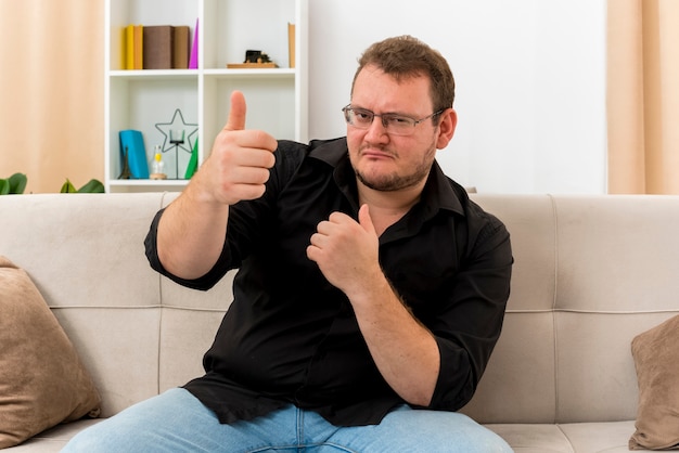 Confident adult slavic man in optical glasses sits on armchair thumbs up with two hands looking at camera inside living room