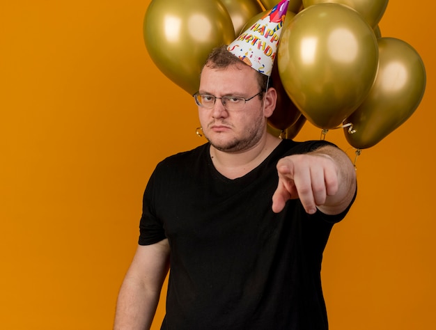Уверенный взрослый славянский мужчина в оптических очках и кепке на день рождения стоит перед гелиевыми шарами, указывая на камеру