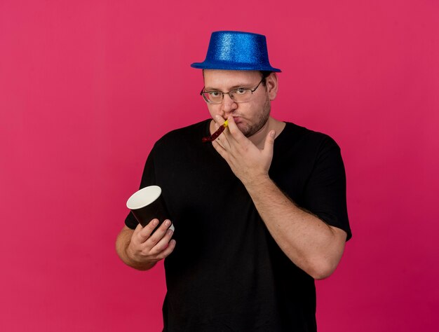 Уверенный взрослый славянский мужчина в оптических очках в синей шляпе держит бумажный стаканчик и дует в свисток