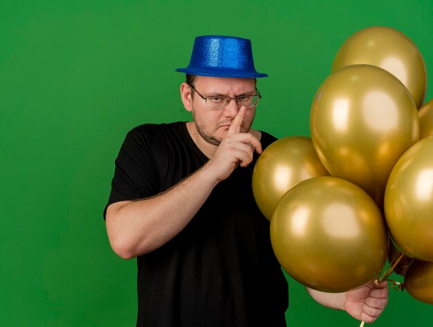 Confident adult slavic man in optical glasses wearing blue party hat holds helium balloons and gestures silence sign 