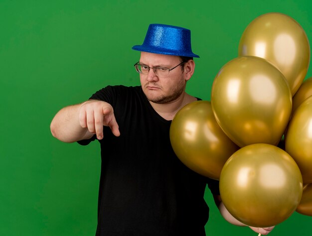 Уверенный взрослый славянский мужчина в оптических очках в синей шляпе держит гелиевые шары и указывает на камеру