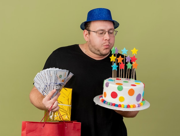 Confident adult slavic man in optical glasses wearing blue party hat holds money gift box paper shopping bag and pretends to blow candles on birthday cake isolated on olive green wall