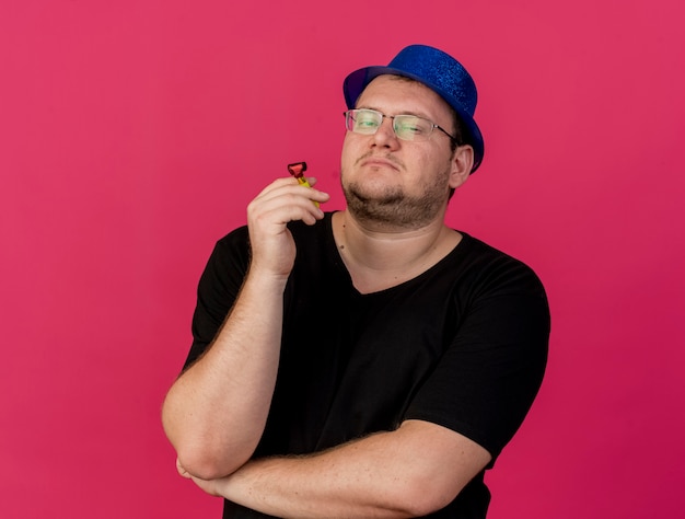 Free photo confident adult slavic man in optical glasses wearing blue party hat holds party whistle