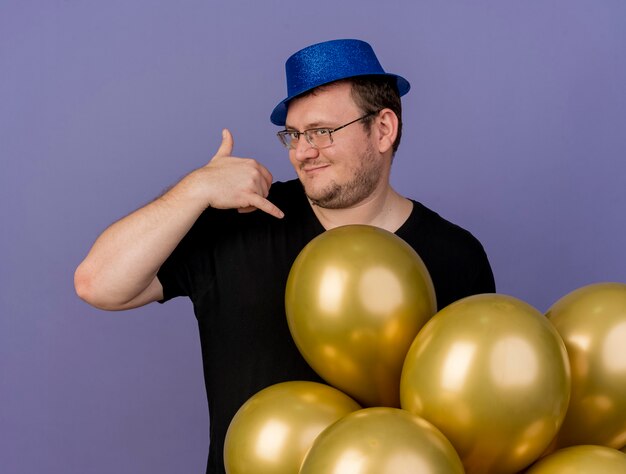 Confident adult slavic man in optical glasses wearing blue party hat stands with helium balloons gesturing call me sign 