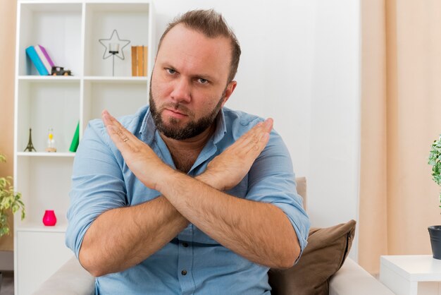 Confident adult slavic man sits on armchair crossing hands gesturing no sign looking at camera inside  living room