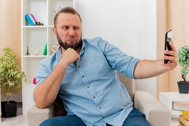 Confident adult slavic man sits on armchair keeping fist looking at phone inside the living room