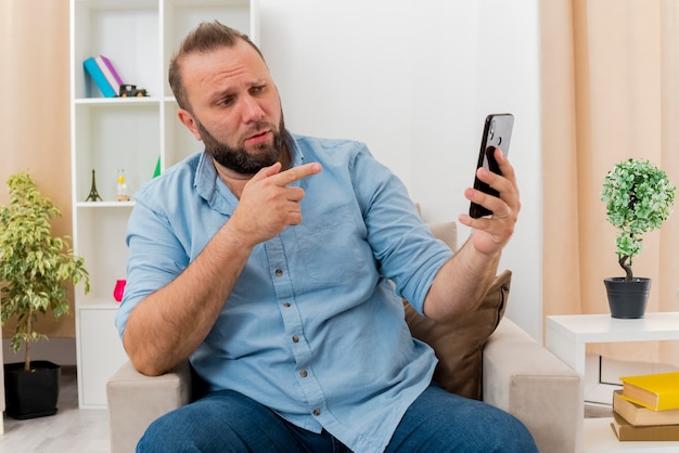 Free photo confident adult slavic man sits on armchair looking and ponting at phone inside the living room