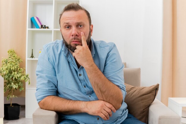 Free photo confident adult slavic man sits on armchair pulling down eyelid inside the living room