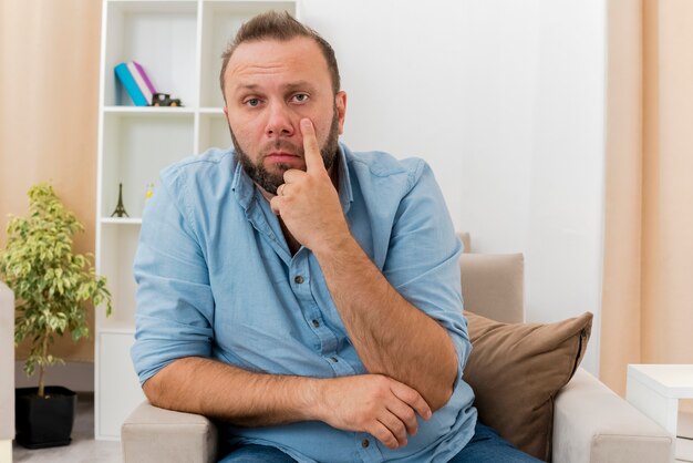 confident adult slavic man sits on armchair pulling down eyelid inside the living room
