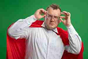 Бесплатное фото Уверенный взрослый славянский супергерой в красной накидке в очках, хватая их, глядя на переднюю часть, изолированную на зеленой стене
