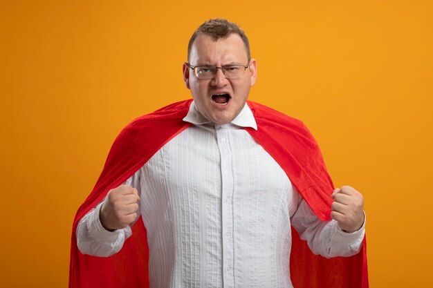 Confident adult slavic superhero man in red cape wearing glasses clenching fists  isolated on orange wall