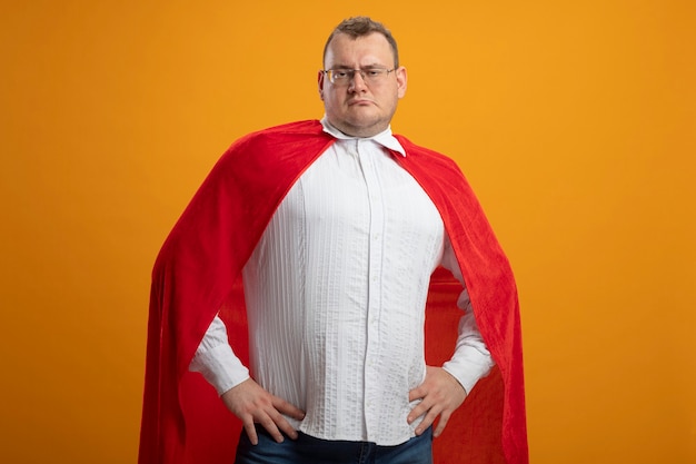 Confident adult slavic superhero man in red cape wearing glasses  keeping hands on waist isolated on orange wall with copy space