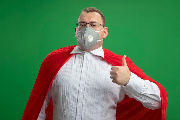 Confident adult slavic superhero man in red cape wearing glasses and protective mask  showing thumb up isolated on green wall