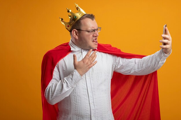 Foto gratuita fiducioso uomo adulto supereroe in mantello rosso con gli occhiali e corona tenendo la mano in aria ammiccante prendendo selfie isolato sulla parete arancione