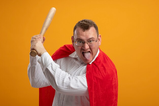 Confident adult superhero man in red cape wearing glasses looking at front showing tongue holding baseball bat getting ready to hit isolated on orange wall