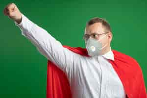 Free photo confident adult superhero man in red cape wearing glasses and protective mask looking at side raising fist up isolated on green wall