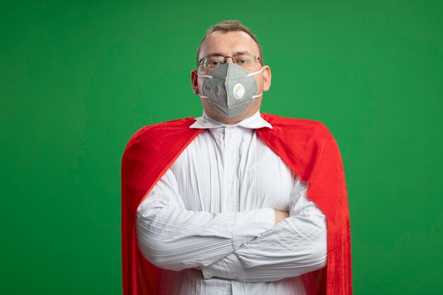 Free photo confident adult superhero man in red cape wearing glasses and protective mask standing with closed posture looking at front isolated on green wall