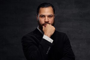 Confident african-american businessman in a black classic jacket, thinking about business while holding hand on chin. isolated on a dark background.