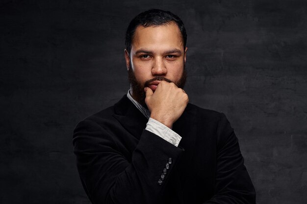 Confident African-American businessman in a black classic jacket, thinking about business while holding hand on chin. Isolated on a dark background.