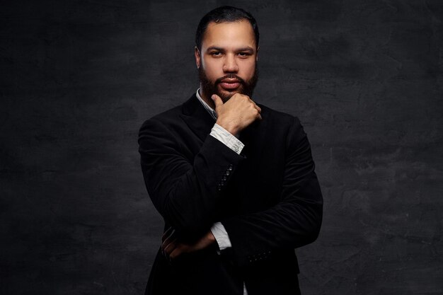 Confident African-American businessman in a black classic jacket, thinking about business while holding hand on chin. Isolated on a dark background.