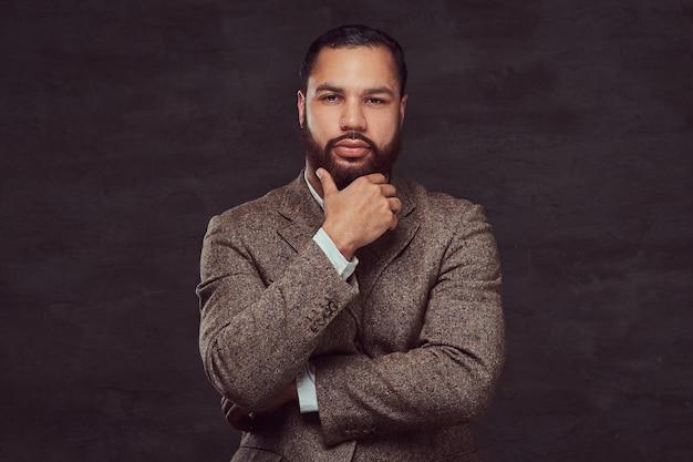 Confident African-American businessman in a brown classic jacket, thinking about business while holding hand on chin. Isolated on a dark background.