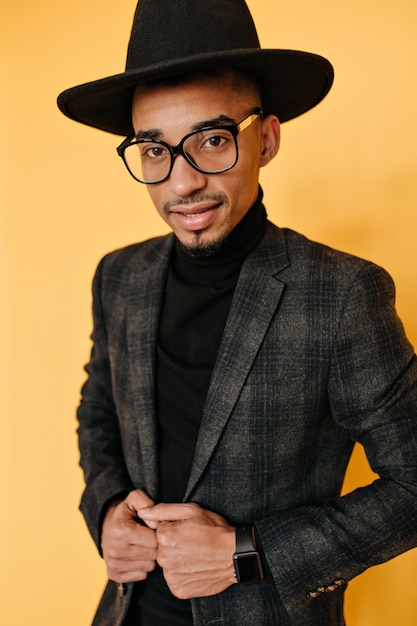 Free photo confident african man in black clothes and trendy wristwatch posing . indoor photo of handsome mulatto guy in glasses isolated on orange wall.