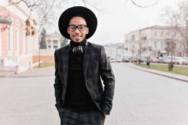 Confident african man standing with hands in pockets and interested face expression. Outdoor photo of gorgeous black male model waiting someone on the street in morning.