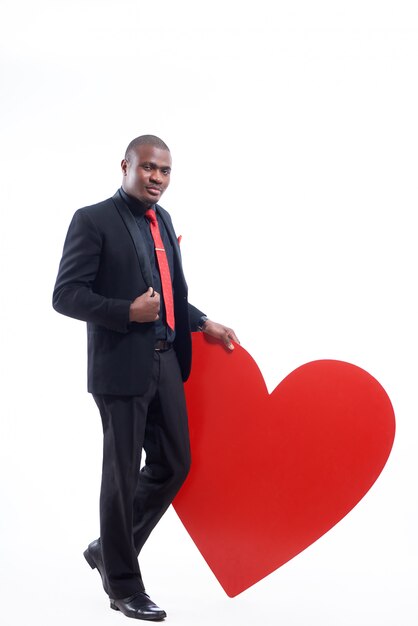 confident african man wearing elegant suite and red tie leaning by hand on big red heart