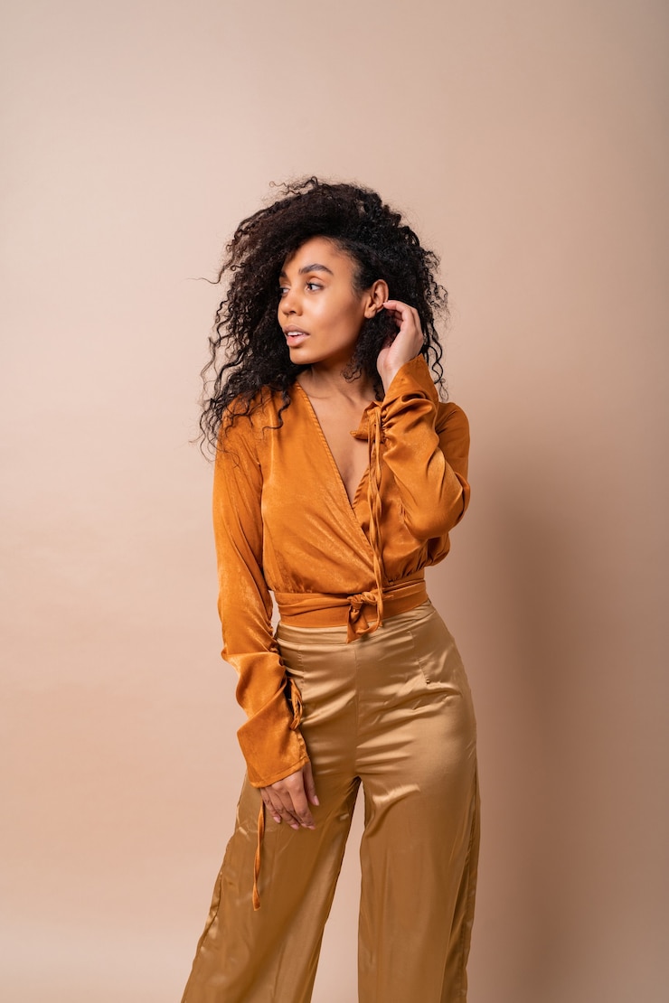 Free Photo confident african woman with perfect curly hairs in casual orange blouse and golden pants posing on beige wall.
