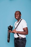 confident and handsome looking young adult person wearing trendy clothes while having photo device. smiling heartily professional photographer having dslr camera while standing on blue background.
