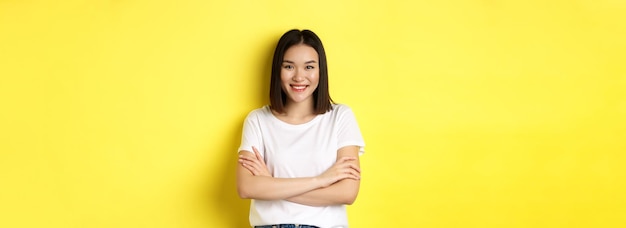 無料写真 自信に満ちたスタイリッシュなアジアの女性が、黄色の背景の上に立ち、胸に腕を組んで微笑む