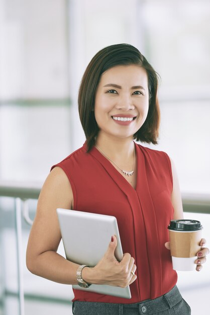 Confident Asian business lady posing indoors on balcony with takeaway coffee and tablet