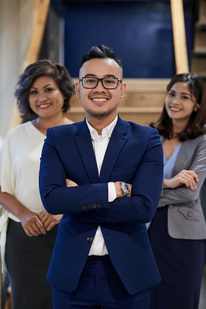 Confident Asian businessman posing with crossed arms, and two female colleagues standing behind
