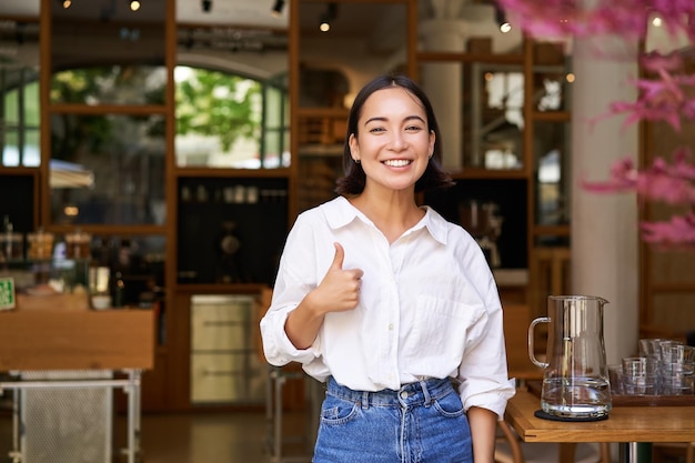 Free photo confident asian businesswoman showing thumbs up standing near entrance of her cafe or restaurant recommending place