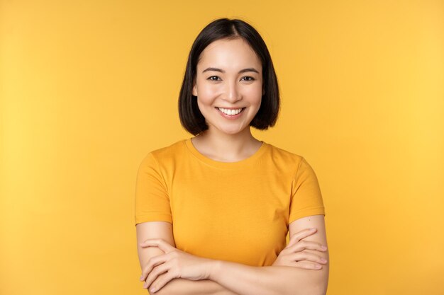 Уверенная азиатская девушка скрещивает руки на груди, улыбаясь и выглядя напористой, стоя на желтом фоне