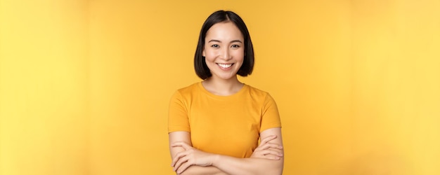 Confident asian girl cross arms on chest smiling and looking assertive standing over yellow background