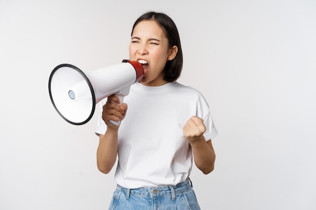 Уверенная азиатка кричит в мегафон, кричит и протестует Девушка-активистка использует динамик, чтобы говорить громче, стоя на белом фоне