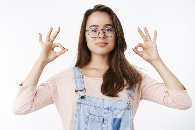 Confident assertive female professional in overalls and glasses assuring work going great, showing okay gesture with self-assured expression, smiling satisfied, liking product.